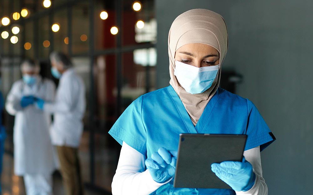 Young Arab woman doctor in hijab, medical mask and gloves standing in hospital and using tablet device. Muslim female medic in traditional headscarf tapping on gadget computer in clinic. Covid-19.